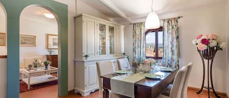 Uma sala de jantar com uma mesa de madeira posta para quatro pessoas, um armário branco com portas de vidro e um arranjo floral sobre um suporte.
