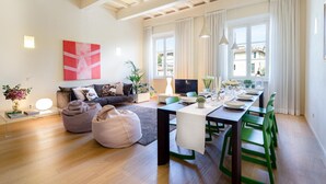 A modern living room with a sofa, a dining table set for four, and a large window with curtains. - Casa Cachi 2 in Lucca (Lucca)