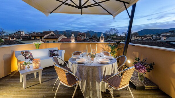 A rooftop terrace with a round table set for dining, surrounded by wicker chairs, under a large umbrella.