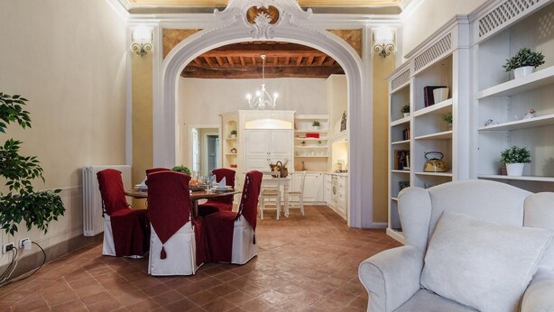 A dining area with a high ceiling, ornate ceiling design, and a chandelier. There is a dining table with chairs, a kitchen in the background, and a white sofa in the foreground. - Casa Daniela in Lucca (Lucca)