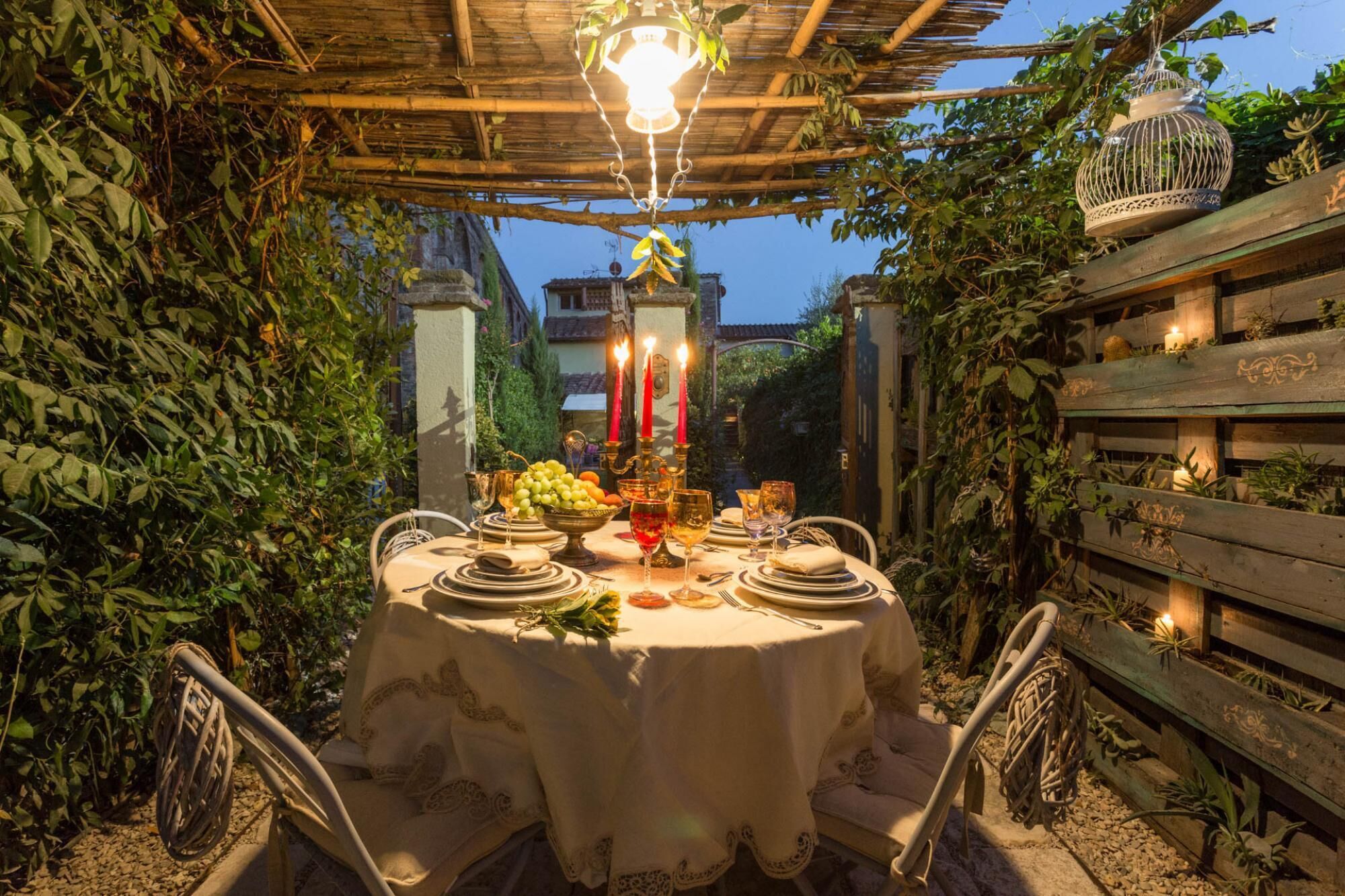 An outdoor dining area with a round table set for a meal, surrounded by greenery and a wooden pergola.