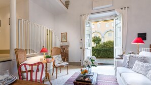 Un salón bien iluminado con un sofá blanco, una mesa de centro de madera y una mesa de comedor con sillas. Se ve una escalera con barandillas blancas y una silla roja. Un ventanal con cortinas blancas da a un patio con vegetación y un edificio al fondo.