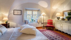Un dormitorio con una cama grande, una ventana con vista a un edificio, un radiador, una silla roja y un aparador con espejo.