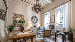 A cozy living room with a wooden dining table, a sofa, and a TV. There's a bookshelf and a large clock on the wall. - Casa Janca in Lucca With 1 Bedrooms and 2 Bathrooms (Lucca)