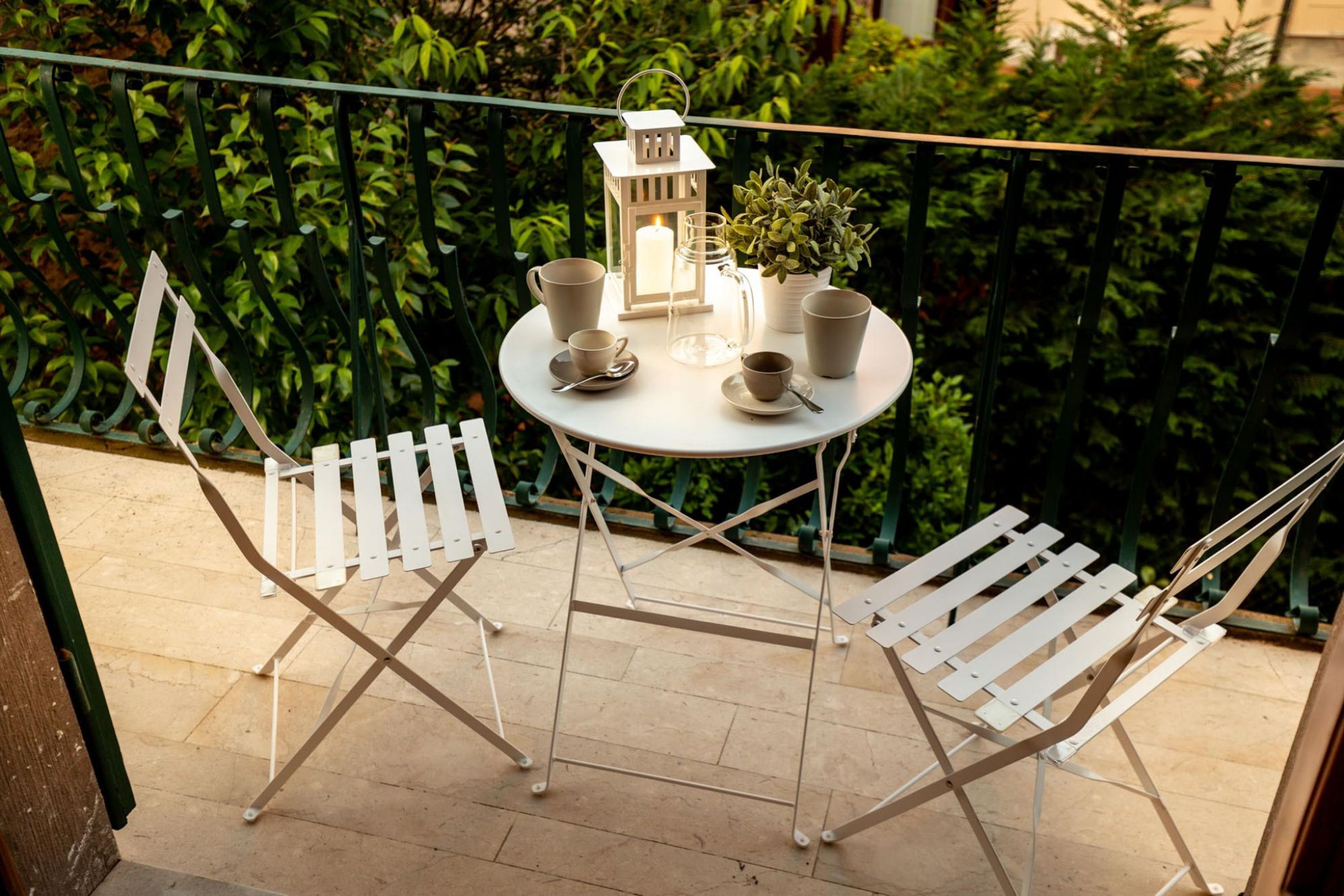 A round table with a white lantern, two white chairs, and a potted plant on a balcony with a green railing.
