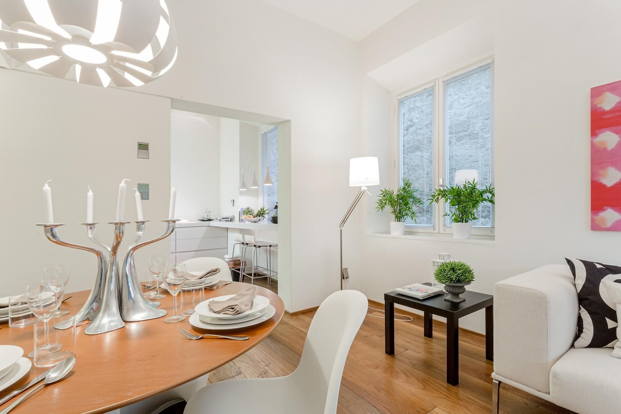 A modern dining area with a wooden table set for four, white chairs, and a chandelier. A kitchen is visible in the background, and there is a window with plants on the windowsill.