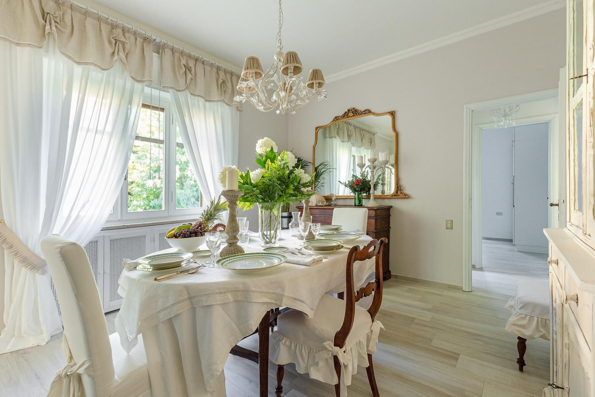 Una sala da pranzo con tavolo apparecchiato per quattro persone, un lampadario, uno specchio e vista sull'esterno.