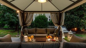 An outdoor seating area with a cushioned bench, wicker furniture, and a table set with candles, fruits, and a bottle. - Villa Olivia in Lucca (Lucca)