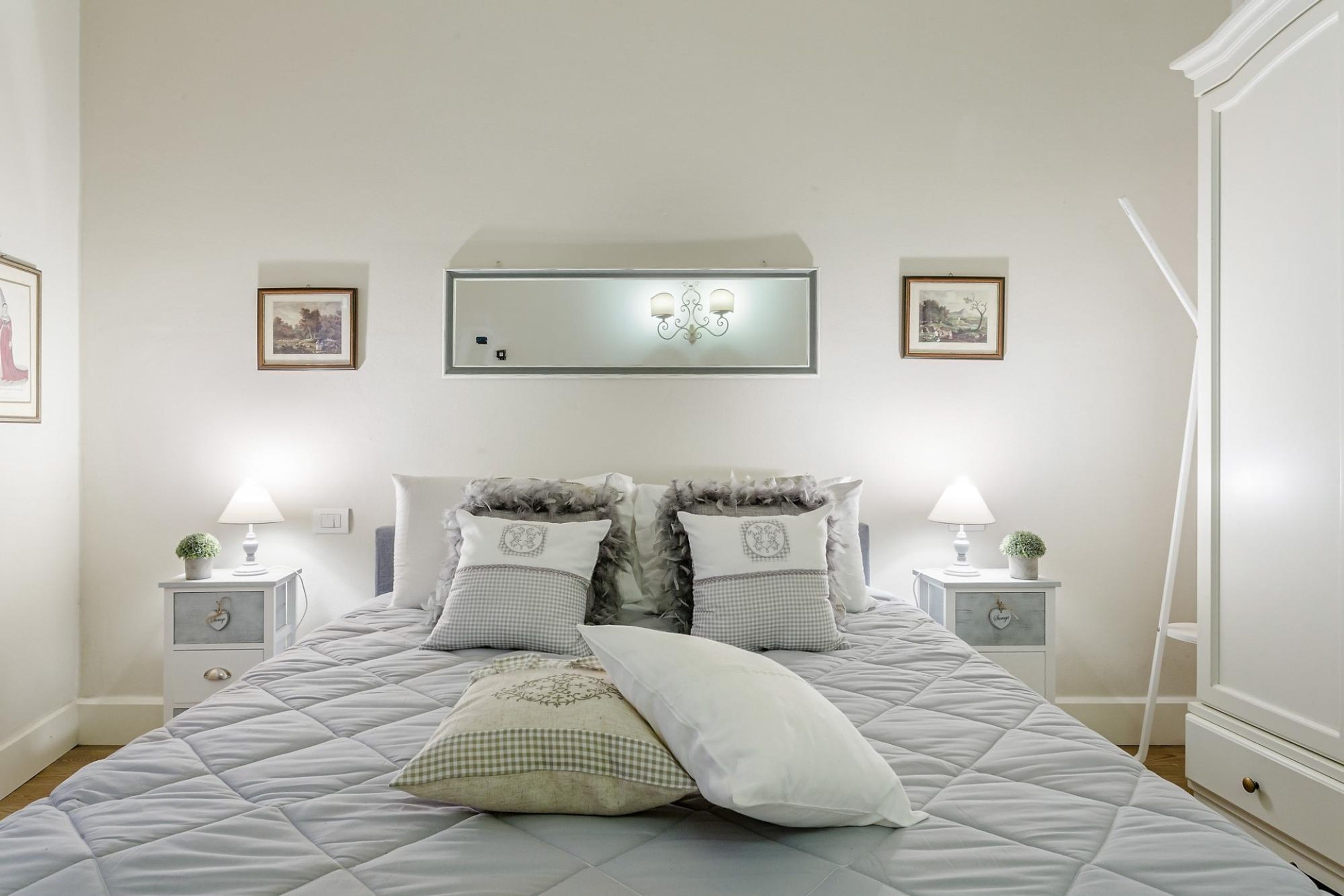 Un letto rifatto con coperta trapuntata e cuscini decorativi, affiancato da comodini con lampade e quadri appesi al muro.