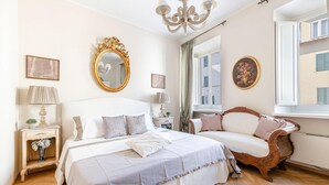 A bedroom with a large bed, a wooden sofa, a chandelier, and a window with a view of buildings. - Casa San Frediano in Lucca (Lucca)