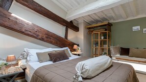 Ein Schlafzimmer mit einem großen Bett, Holzbalken, einer Sitzbank an einem Fenster und einem kleinen Tisch mit einer Lampe.
