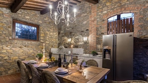 A rustic dining area with a stone wall, wooden ceiling, and a chandelier. A wooden table is set with plates, glasses, and a bottle of wine. - Villa Clara in Capannori (Capannori)