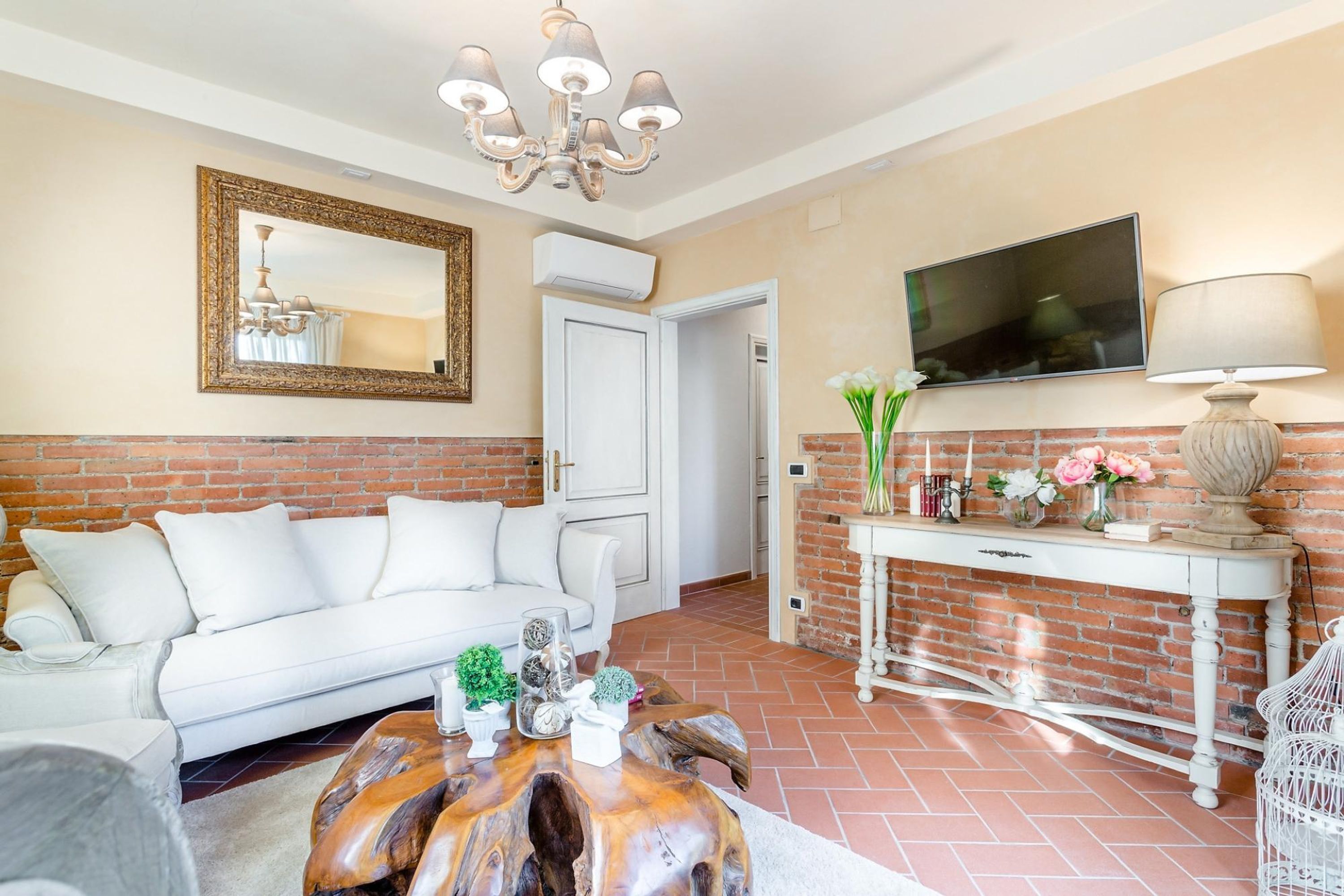 Ein Wohnzimmer mit weißem Sofa, einem Holztisch, einem an der Wand befestigten Fernseher und einem Kamin aus Ziegelsteinen.