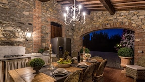 An outdoor dining area with a stone wall, wooden table, wicker chairs, and a chandelier. - Villa Clara in Capannori (Capannori)