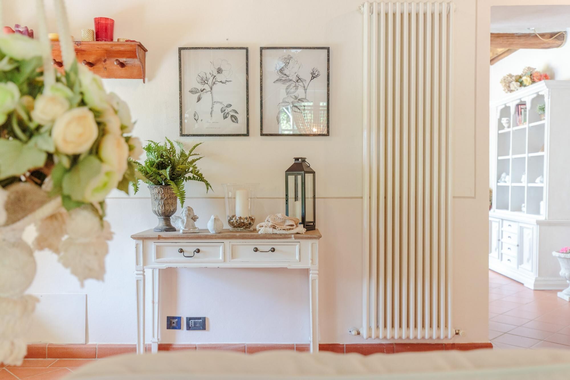 Um quarto com um radiador branco, uma mesa consola branca com objetos decorativos, e duas gravuras botânicas emolduradas na parede.