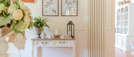 Um quarto com um radiador branco, uma mesa consola branca com objetos decorativos, e duas gravuras botânicas emolduradas na parede.