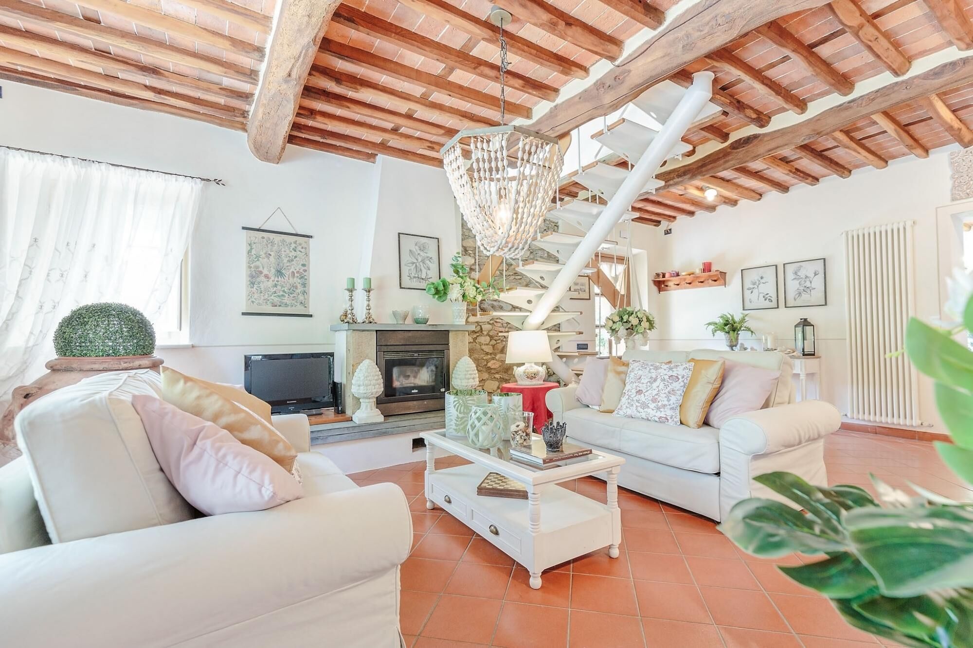 A spacious living room with a fireplace, white sofas, a coffee table, and a staircase with a wooden ceiling.