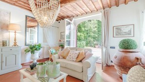 A cozy living room with a white sofa, a hanging light fixture, and a view of greenery outside. - Villa Coccinelle in Pieve di Compito (Capannori)