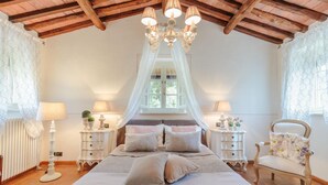 A bedroom with a large bed, a chandelier, bedside tables, a chair, and a window with curtains. - Villa Coccinelle in Pieve di Compito (Capannori)