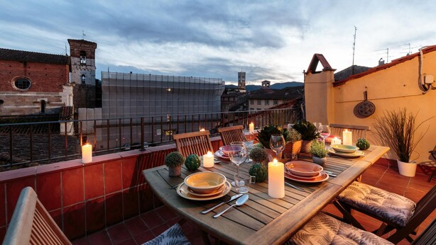Terraço no telhado com mesa de madeira posta para refeições, rodeada por cadeiras, velas e plantas em vasos.