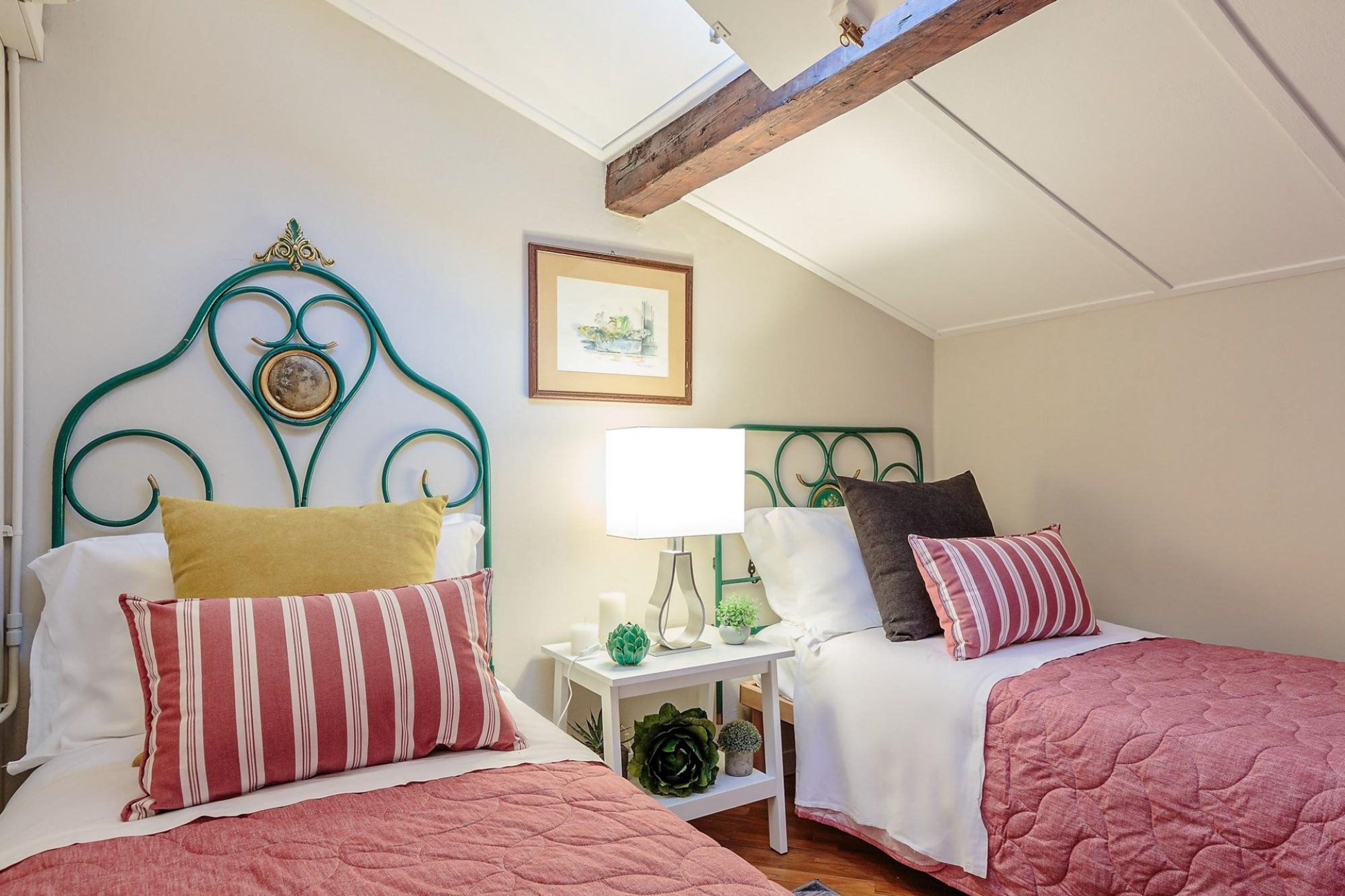 A bedroom with two beds, each with a distinct headboard design, a bedside table with a lamp, and a framed picture on the wall.