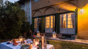 An outdoor dining area with a table set for a meal, surrounded by greenery and a building with arched windows. - Villa Gattaiola in Gattaiola (Lucca)
