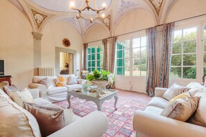 A spacious living room with a large window, a chandelier, and a decorative ceiling. - Villa Gattaiola in Gattaiola (Lucca)