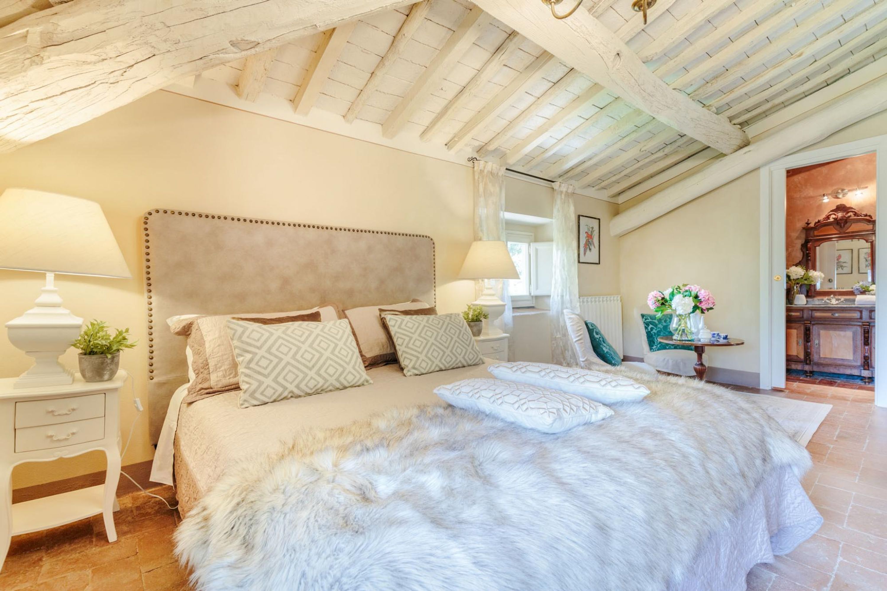 A bedroom with a large bed, two bedside tables, a nightstand, a fluffy white rug, and a wooden ceiling.