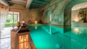 A spacious indoor swimming pool with clear water, wooden beams on the ceiling, and a stone wall with an arched doorway. - Villa Il Palagio in Capannori (Capannori)