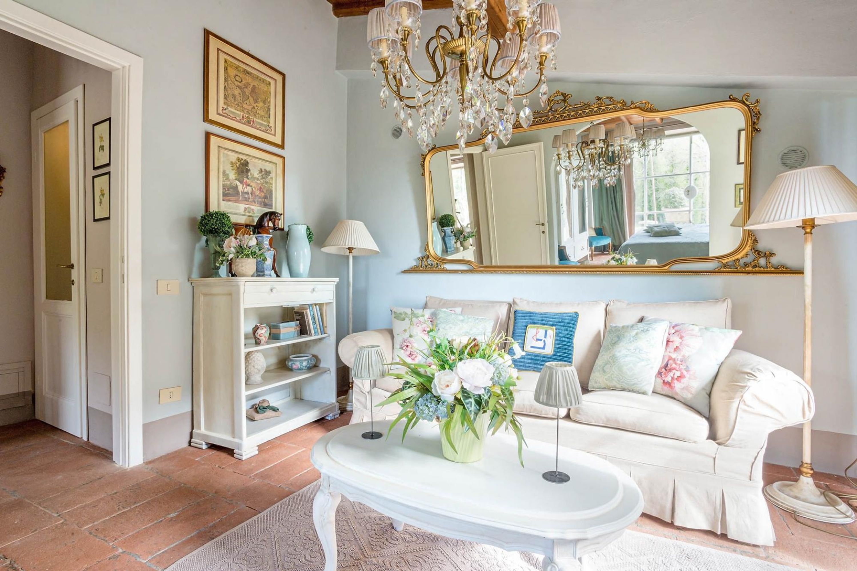 A cozy living room with a white sofa, a coffee table with a floral arrangement, a large mirror, and a chandelier.