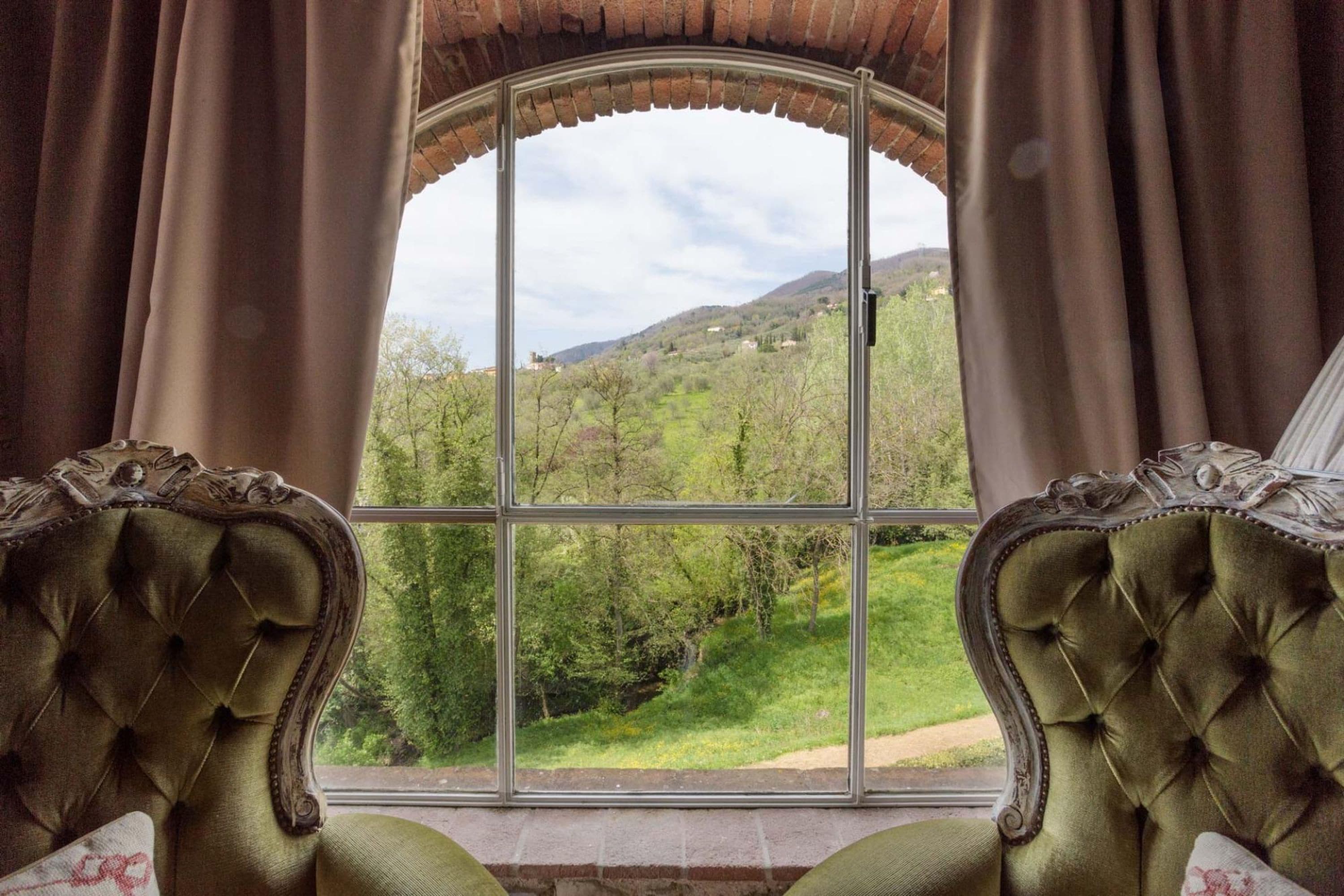 A room with a large window offering a view of a green landscape and hills. There are two upholstered chairs with ornate detailing.