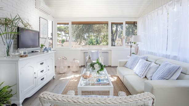 A bright living room with a white sofa, a coffee table with decorative items, a television on a white cabinet, and large windows with a view of trees. - Villa Marygrace in Forte dei Marmi (Forte dei Marmi)