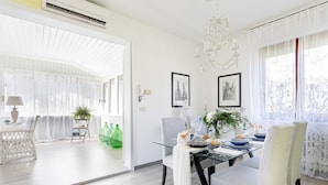 A dining area with a glass table, white chairs, and a chandelier. There are framed pictures on the wall and a plant on the table. - Villa Marygrace in Forte dei Marmi (Forte dei Marmi)