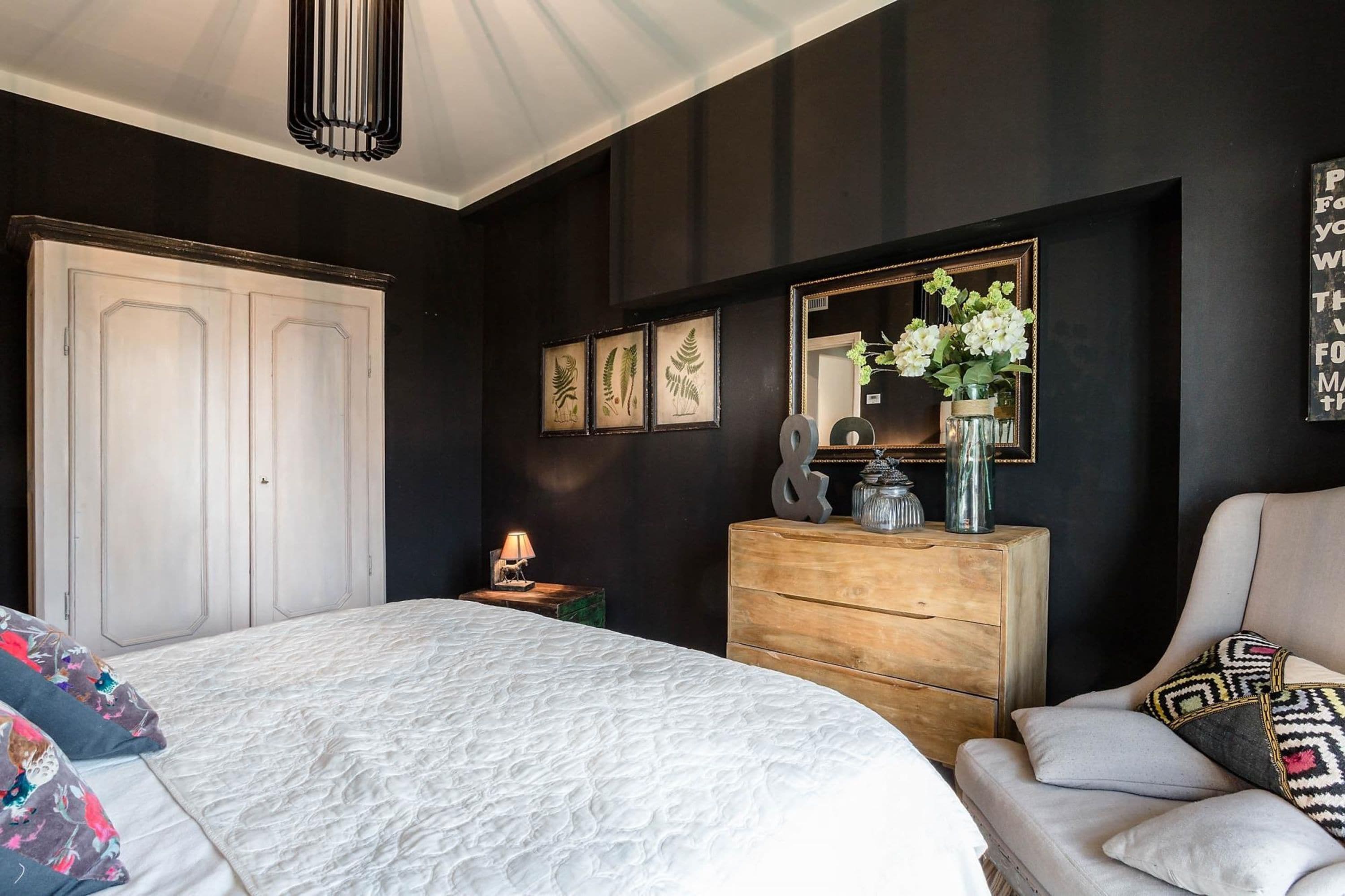 A bedroom with a bed, dresser, chair, and a mirror with a flower arrangement.