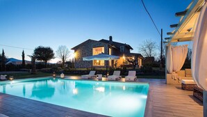 A swimming pool with lounge chairs, a stone house with a lit interior, and a covered outdoor seating area. - Villa Otto in Orentano (Castelfranco di Sotto)