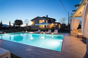 Piscina com espreguiçadeiras, casa de pedra com interior iluminado e área de estar coberta ao ar livre.