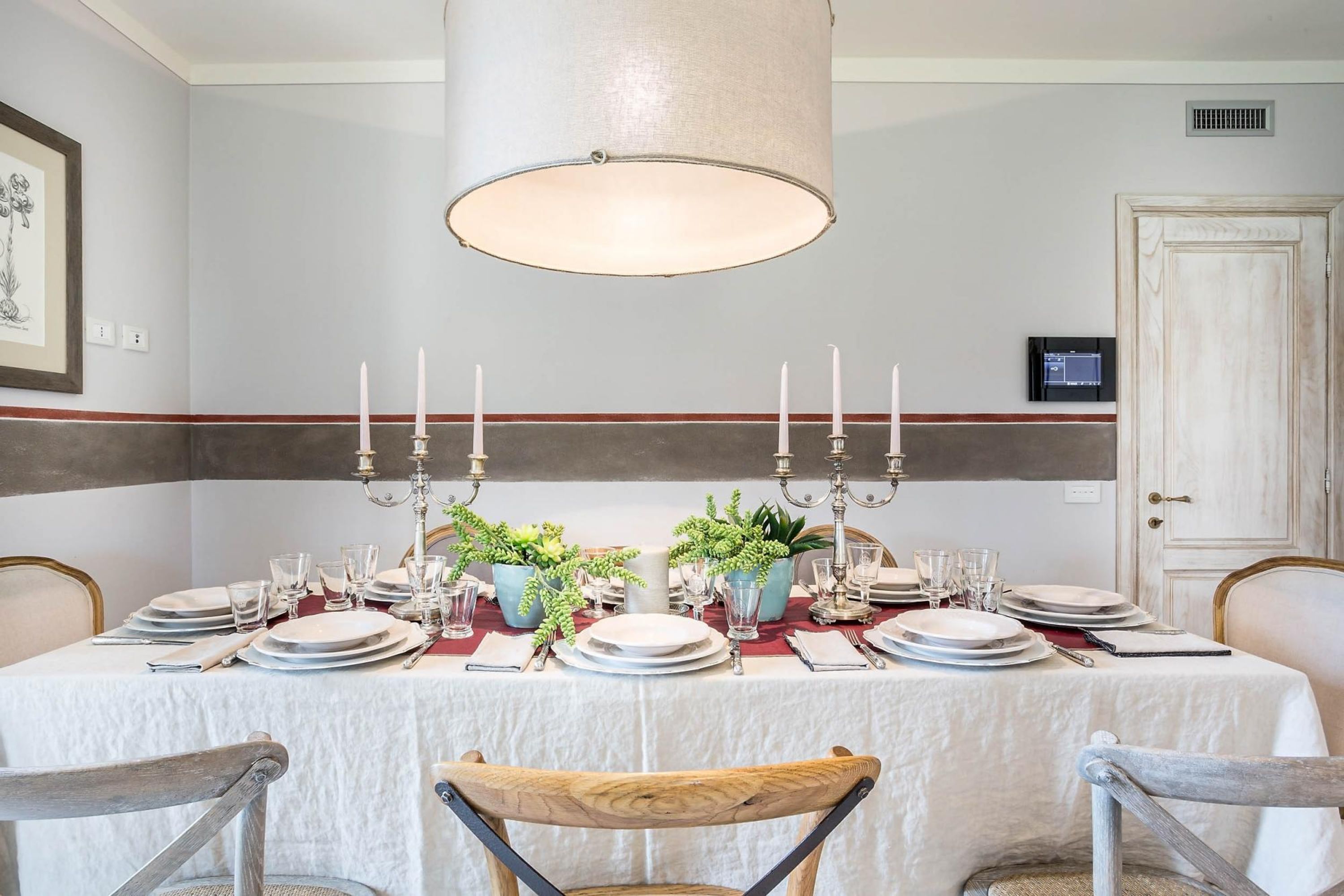 A dining room with a table set for six, featuring a white tablecloth, plates, glasses, and candelabras. There are two potted plants on the table, and the room has a neutral color scheme with a striped wall.