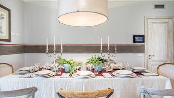 A dining room with a table set for six, featuring a white tablecloth, plates, glasses, and candelabras. There are two potted plants on the table, and the room has a neutral color scheme with a striped wall.
