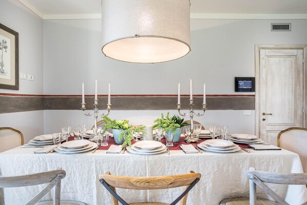 A dining room with a table set for six, featuring a white tablecloth, plates, glasses, and candelabras. There are two potted plants on the table, and the room has a neutral color scheme with a striped wall. - Villa Otto in Orentano (Castelfranco di Sotto)