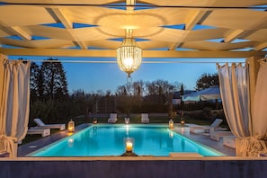 Piscina com espreguiçadeiras e velas, sob uma pérgola com cortinas.