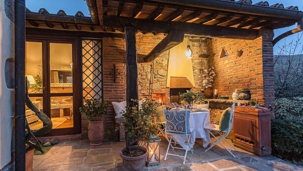 A cozy outdoor patio with a stone floor, a table set for dining, and a brick wall with a stone fireplace. - Rosyabate Cottage in Uzzano (Uzzano)