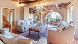 A spacious living room with a high ceiling, exposed wooden beams, and a chandelier. There are two sofas, a coffee table, and a fireplace.