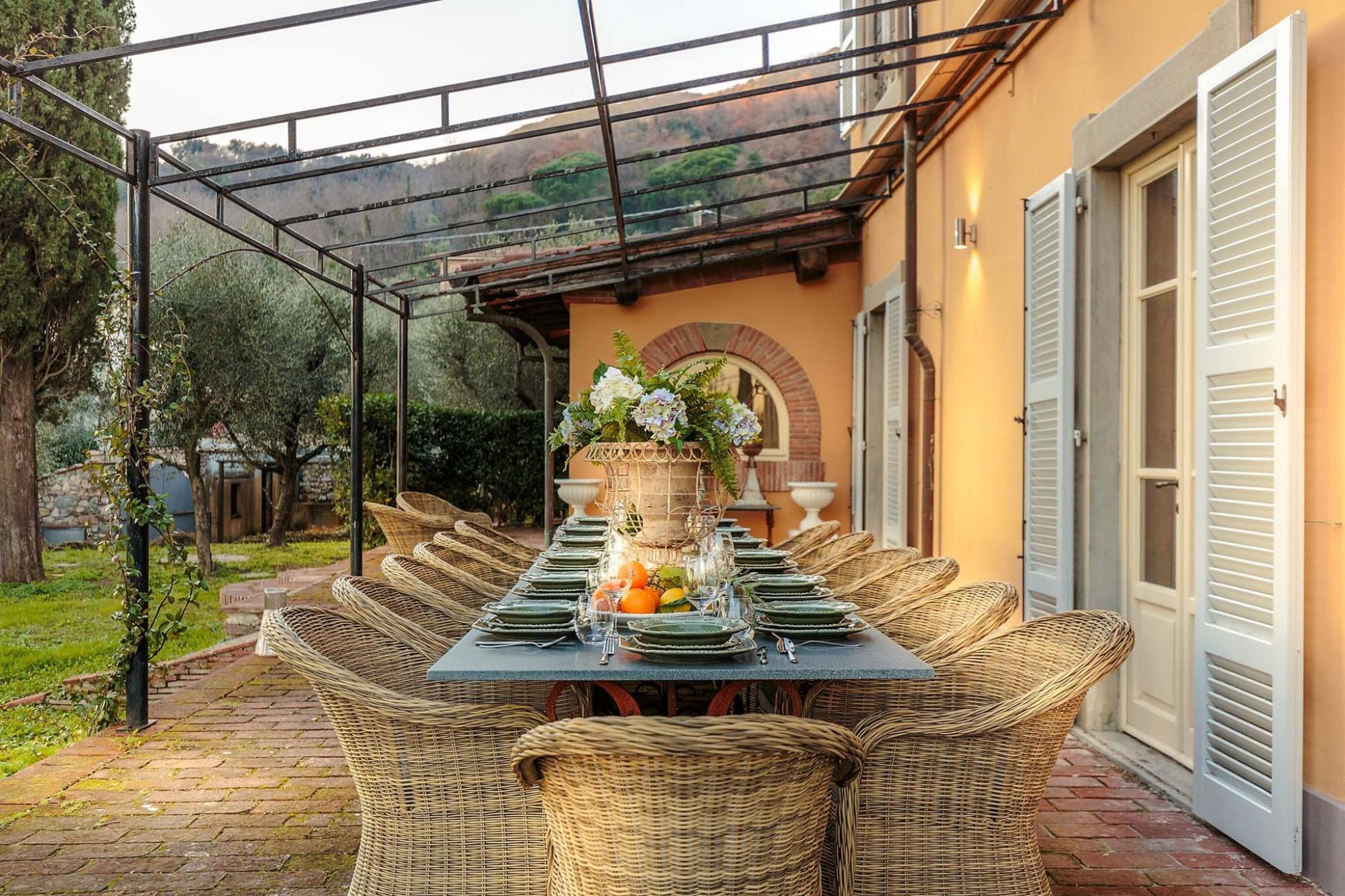 Un área de comedor al aire libre techada, con sillas de mimbre y una mesa dispuesta para una comida, con un centro de mesa floral y fruta.