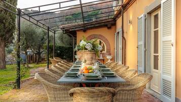 Un área de comedor al aire libre techada, con sillas de mimbre y una mesa dispuesta para una comida, con un centro de mesa floral y fruta.