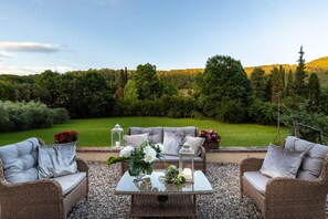 Een terras met rieten meubilair en een glazen tafel, omringd door een weelderige tuin en uitzicht op een beboste helling.