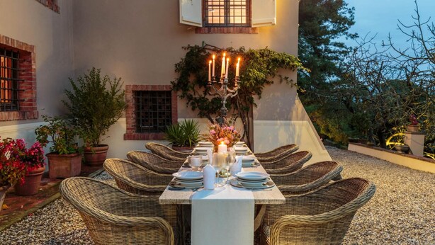 An outdoor dining area with wicker chairs and a table set for six, surrounded by potted plants and a building with a brick facade. - Villa Viola Residenze Seicento (Capannori)