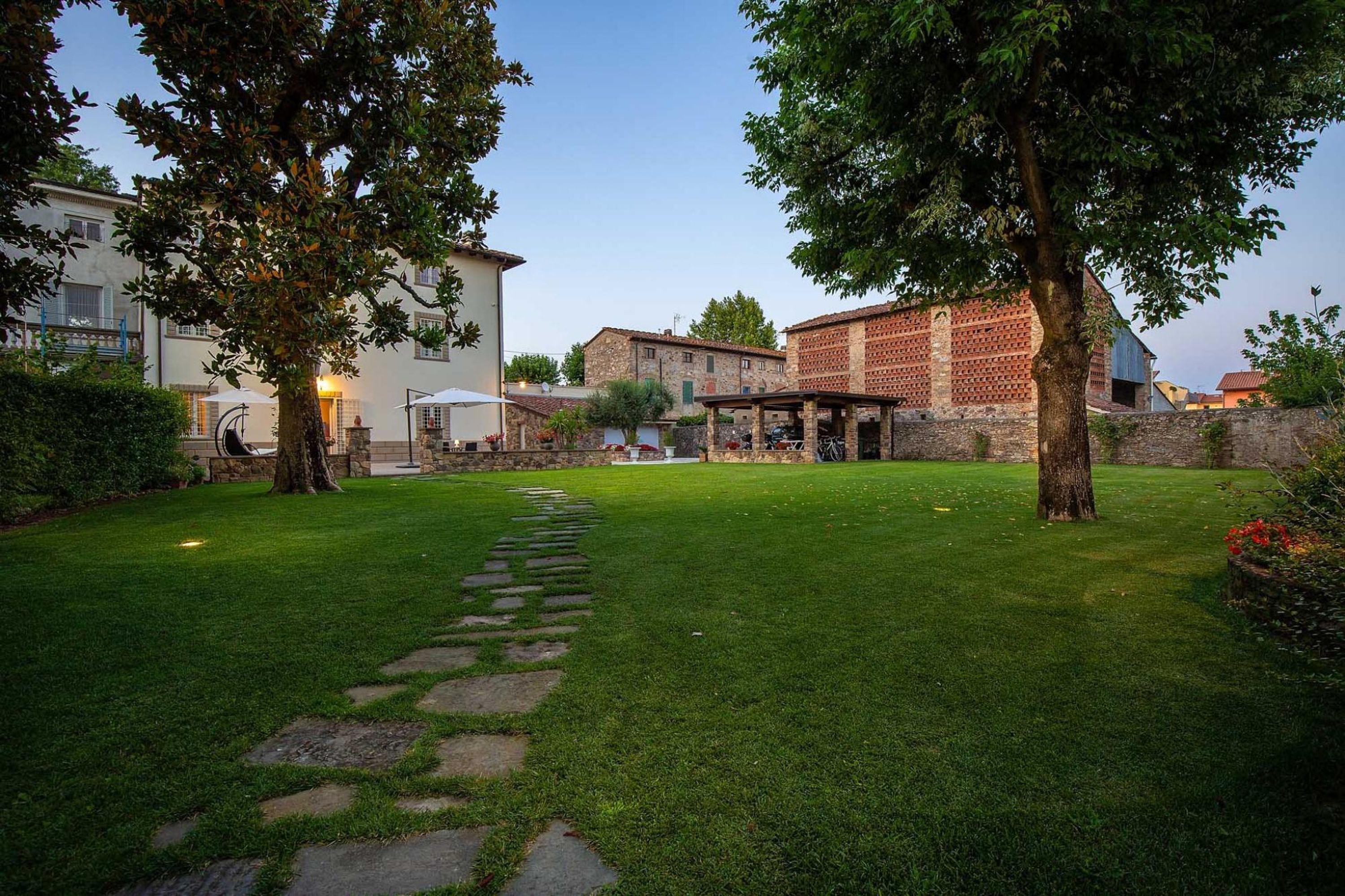 Un giardino curato con un vialetto in pietra che conduce a un'area salotto, circondato da alberi e edifici.