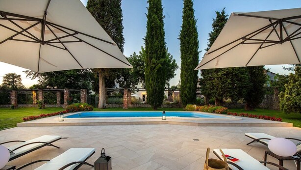 Un espace piscine extérieur doté de chaises longues et de parasols, entouré d’un jardin bien entretenu et de grands arbres.