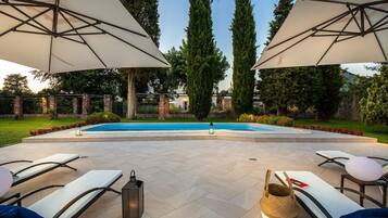 Area piscina all'aperto con lettini e ombrelloni, circondata da un giardino curato e alberi alti.