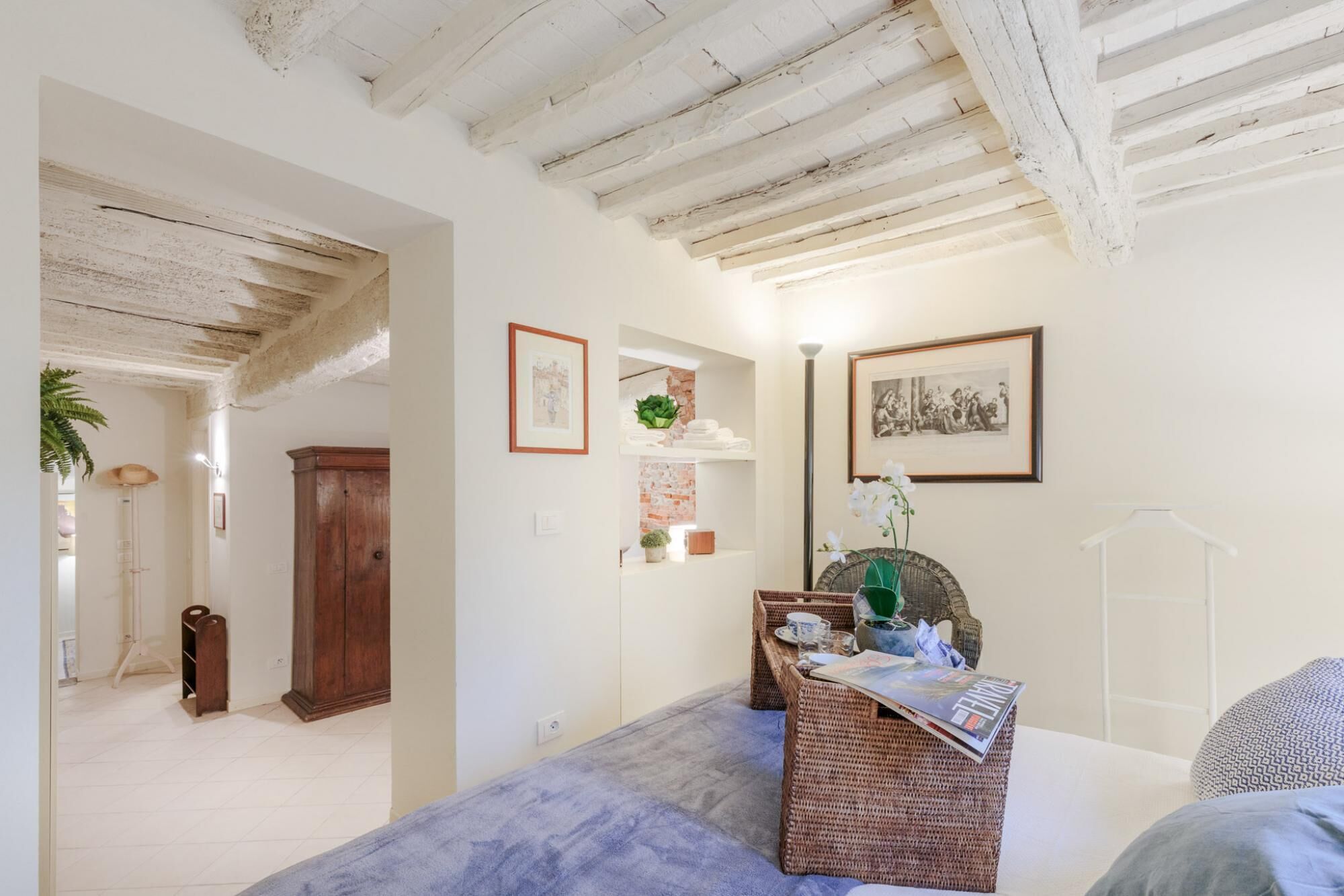 Een slaapkamer met een bed, een rieten bijzettafeltje, een wandplank met decoratieve objecten en een deur naar een andere kamer.